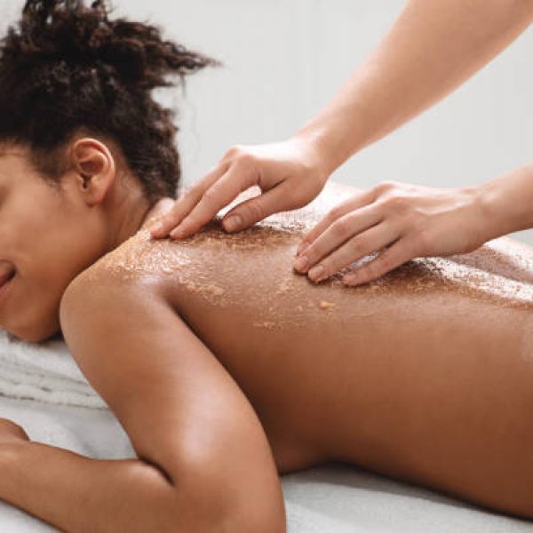 Spa therapist scrubbing black woman skin, using natural beauty products for spa, closeup